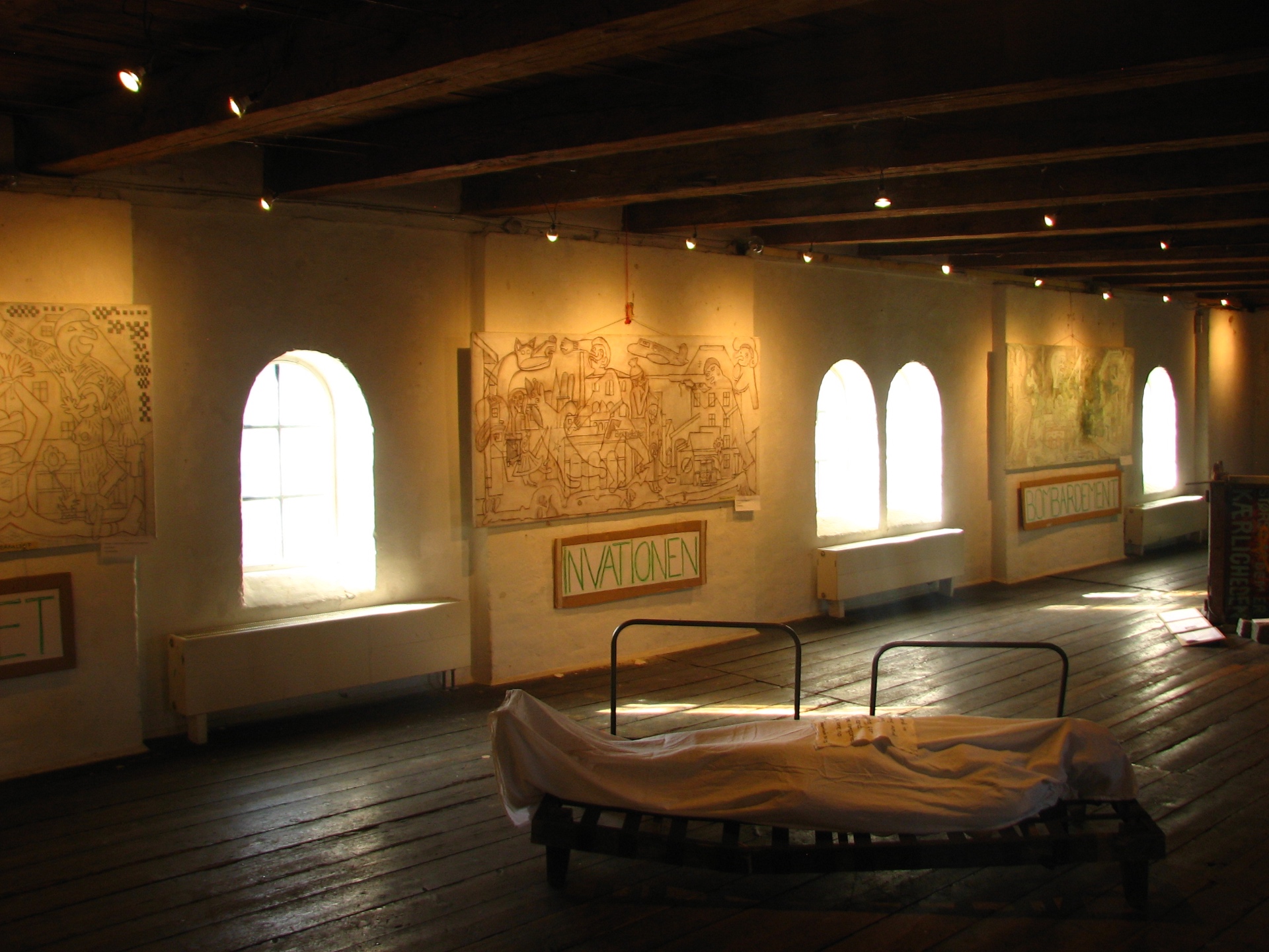 A long timber-beamed hall in a former military barracks, with arched windows along one wall and large drawings displayed between them.