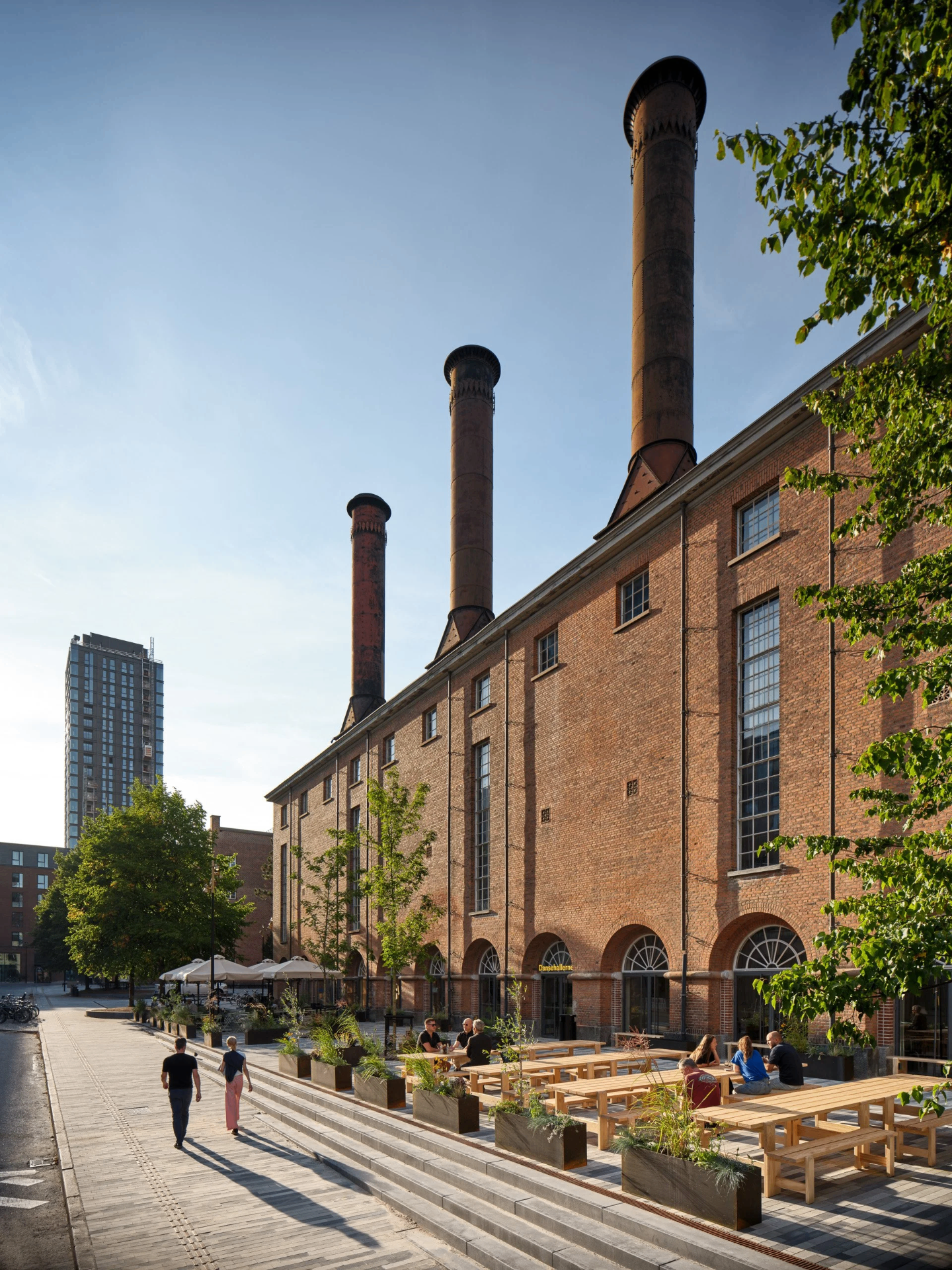 Brick facade of Dansehallerne with three tall industrial chimneys against a clear sky, wooden benches and planters in front