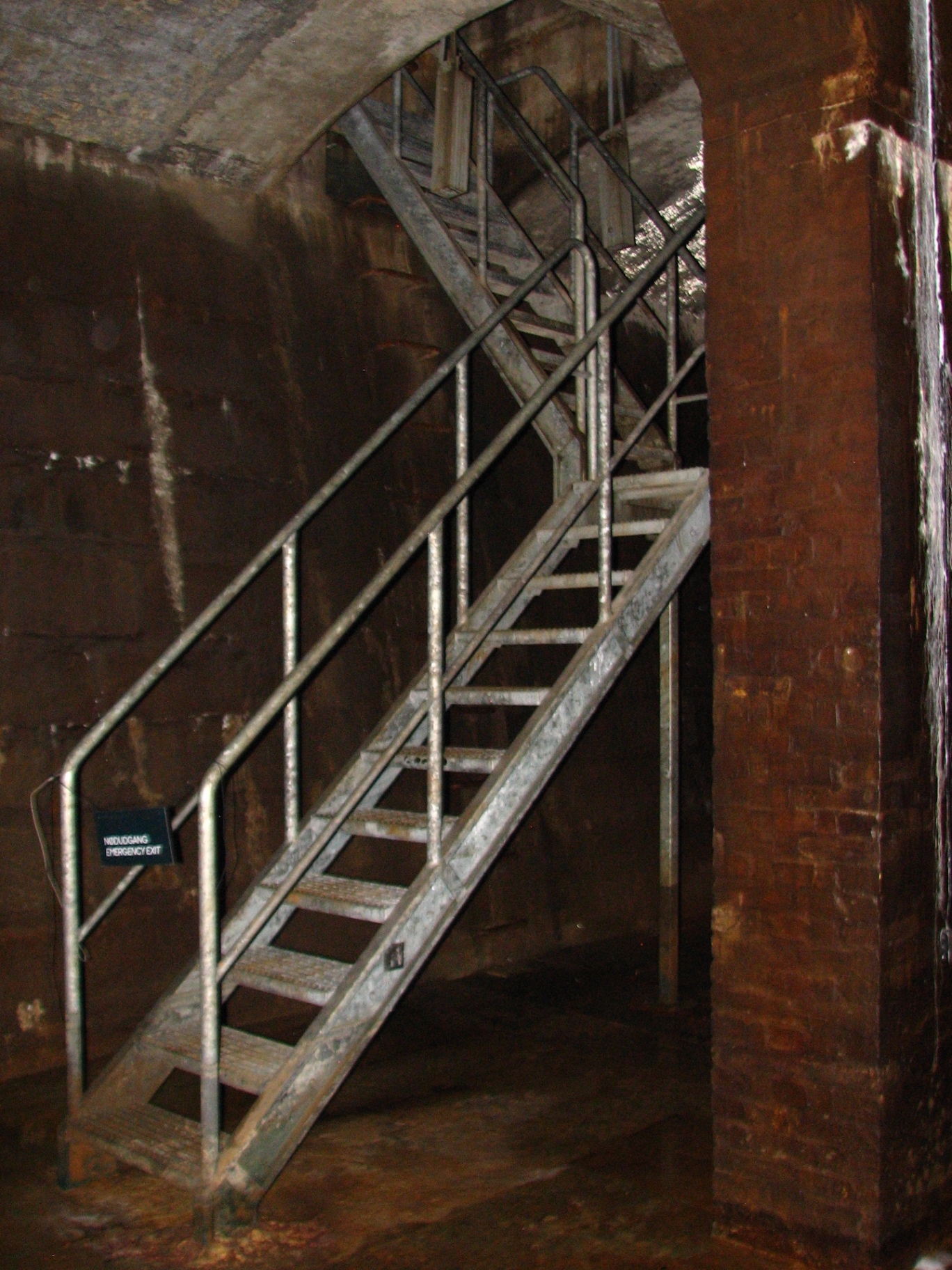Metal staircase marked Nødudgang / Emergency Exit rising through the damp brickwork of the cistern.