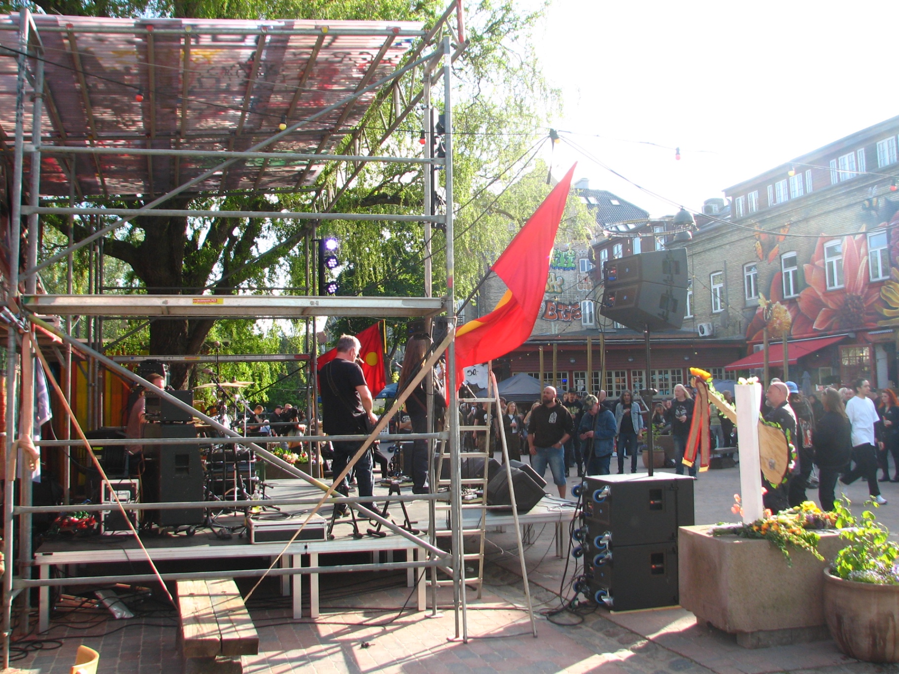 An outdoor concert stage built from scaffolding, with red flags and a crowd gathering around it under tall trees.