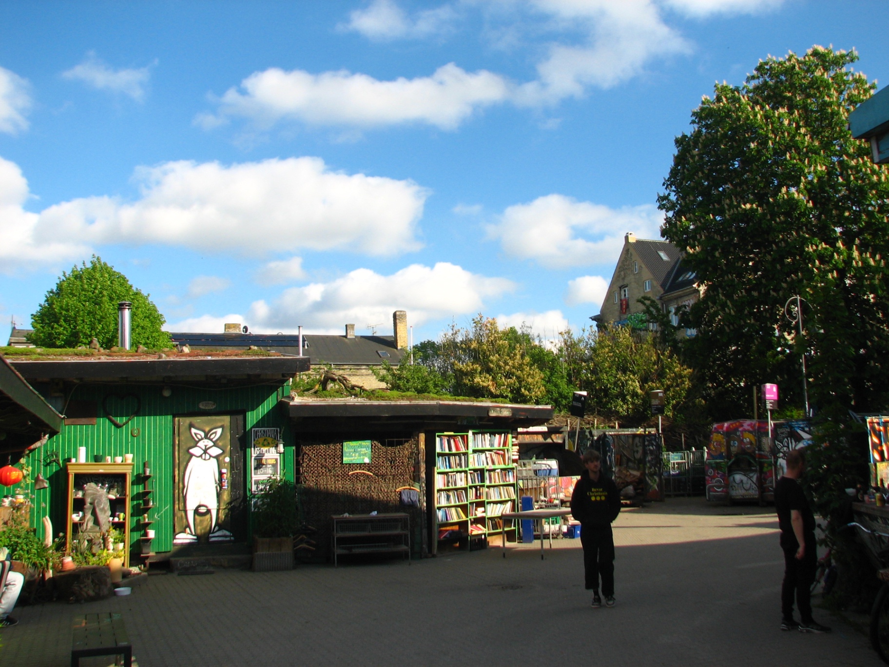 A small square in Christiania with a green-painted shed, a turf-roofed shelter and open bookshelves of second-hand books.