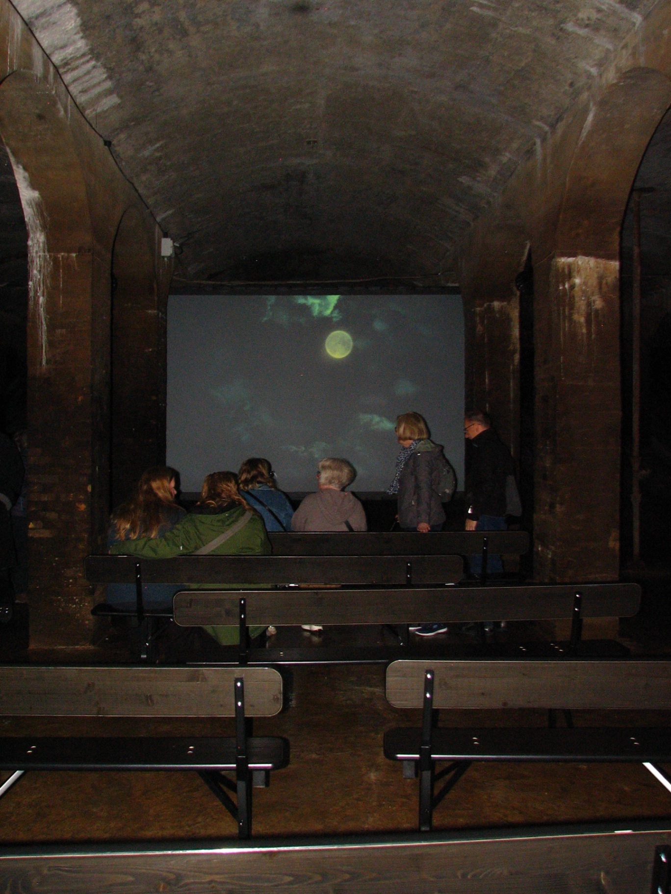 Visitors seated on wooden benches inside Cisternerne, watching a projected moon glow on a screen between concrete pillars.