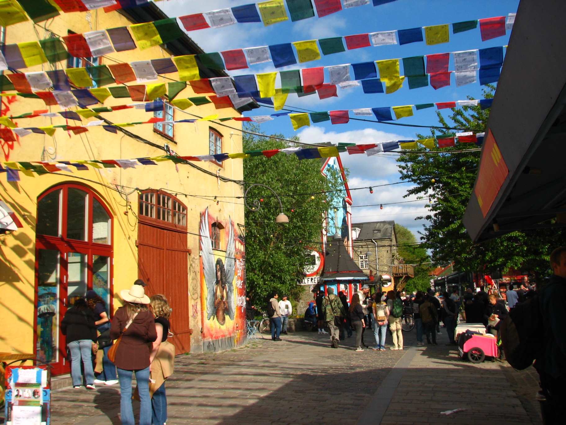A sunlit lane in Christiania crowded with people, criss-crossed overhead with Tibetan prayer flags, a yellow building and street art on either side.